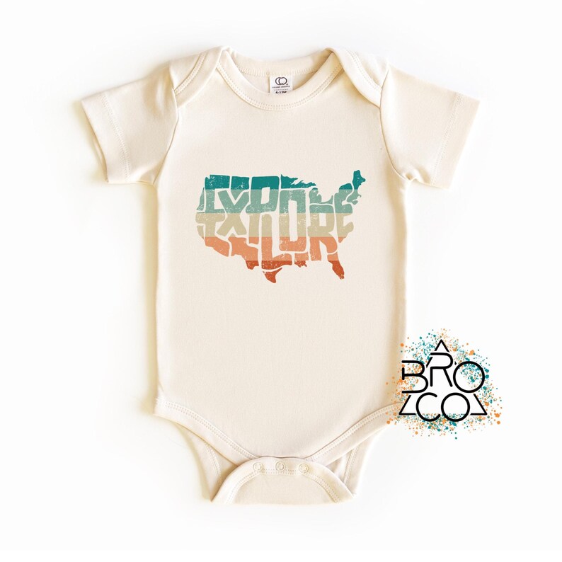 May include: A cream-colored short-sleeve baby bodysuit featuring a distressed graphic of the United States map filled with the word EXPLORE in horizontal stripes of teal, beige, and orange. A logo with the text BRO CO is visible.