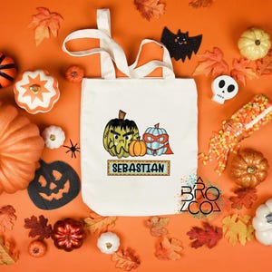Puede incluir: Un bolso tote crema con calabazas de dibujos animados y el nombre "SEBASTIAN". La imagen est&aacute; rodeada de calabazas naranjas, hojas de oto&ntilde;o, una calavera, un murci&eacute;lago y caramelos sobre un fondo naranja.