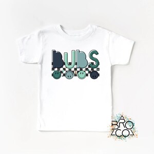 May include: White t-shirt with the word "BUBS" in green and blue letters with a checkered pattern and smiley faces.