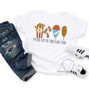 May include: White t-shirt featuring a graphic of popcorn, peanuts, a snow cone, and a corn dog. The text on the shirt reads "I'm here for the concession stand."