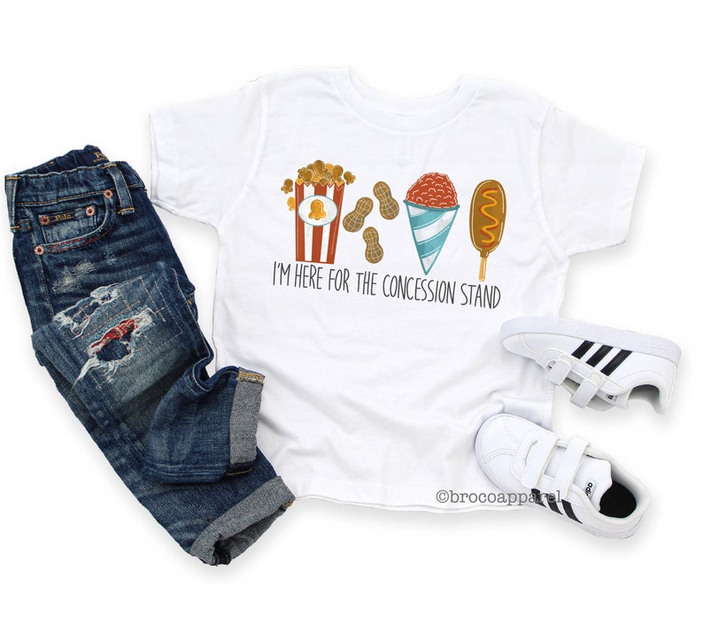 May include: White t-shirt featuring a graphic of popcorn, peanuts, a snow cone, and a corn dog. The text on the shirt reads "I'm here for the concession stand."
