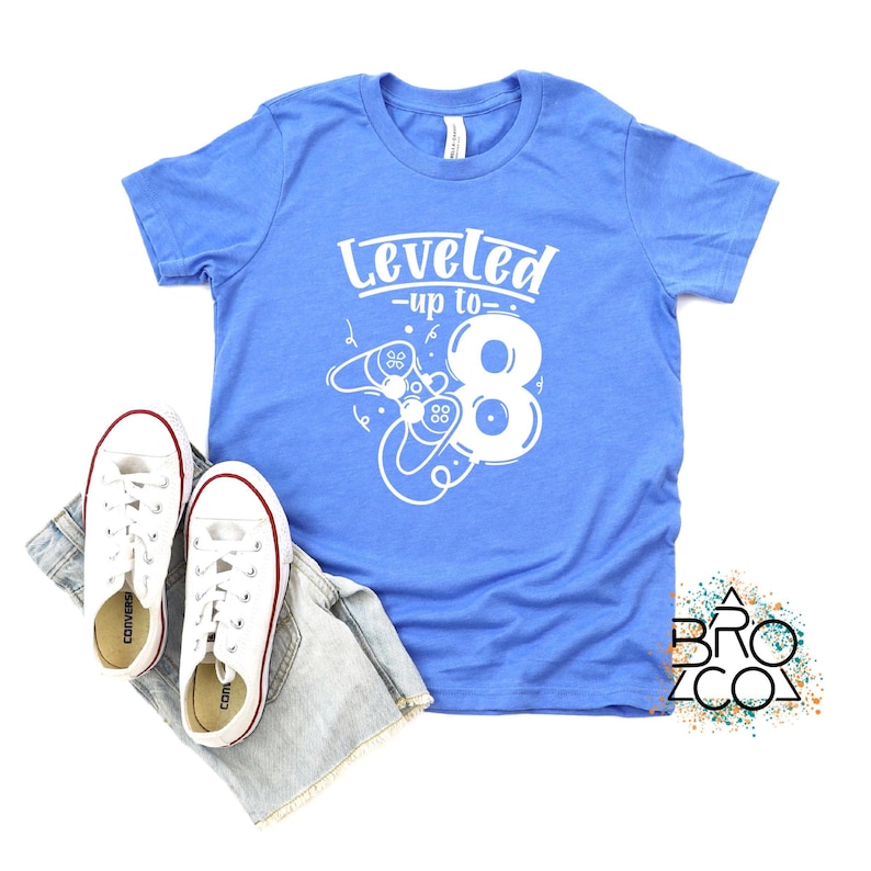 May include: A cornflower blue t-shirt with the text "Leveled up to 8" in white, featuring a video game controller design. The shirt is paired with white trainers and denim shorts, ideal for a birthday celebration.