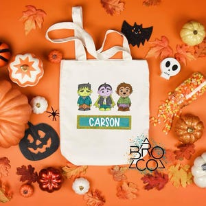 Puede incluir: Un bolso tote crema con monstruos de dibujos animados: Frankenstein, un vampiro y un hombre lobo. El nombre "CARSON" est&aacute; en una pancarta turquesa. Decoraciones de Halloween, calabazas y hojas.