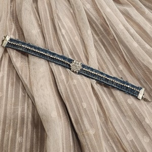 May include: A denim bracelet with a silver rhinestone embellishment. The bracelet is on a light brown tulle fabric.