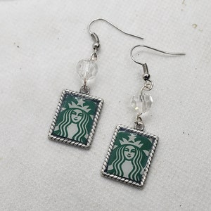 May include: A pair of silver earrings with a green Starbucks logo charm and a clear crystal bead on each earring.