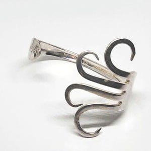 May include: A silver-toned metal bracelet crafted from a repurposed fork. The handle of the fork forms the band of the bracelet, while the tines are curved and spread outward in a fan-like arrangement, creating a distinct open-ended design.