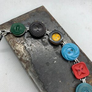 May include: A bracelet made of silver chain with colorful buttons. The buttons are green, black, yellow, blue, red, and blue. The bracelet is on a gray metal surface.