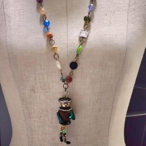 May include: A colorful beaded necklace with a unique pendant featuring a brown figure with green accents. The pendant is made of wood and has a beaded tassel.