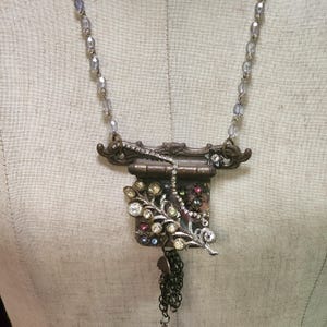 May include: A vintage-inspired necklace with a brown metal pendant adorned with rhinestones and a dangling chain. The pendant features a small, ornate frame with a hinged door and a floral design.