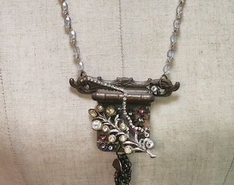 May include: A vintage-inspired necklace with a brown metal pendant adorned with rhinestones and a dangling chain. The pendant features a small, ornate frame with a hinged door and a floral design.