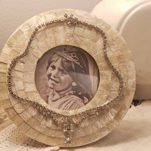 May include: A circular picture frame crafted from iridescent white shell tiles, featuring a central black and white photograph of a child wearing a tiara. A silver-toned rhinestone necklace is draped over the frame, secured at the top.