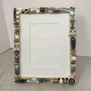 May include: A rectangular photo frame with a gold-colored metal frame. The frame is decorated with an assortment of colorful stones, shells, and decorative elements. The frame has a white interior.