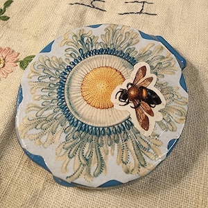 May include: A round, blue and white ceramic trinket box with a floral design and a bee cutout. The bee is brown and yellow with black stripes.