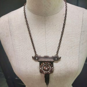 May include: A brown chain necklace with a pendant featuring a rectangular frame with a decorative design of rhinestones and a tassel.