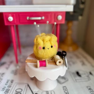 May include: A whimsical yellow felt pincushion with a smiling face and beaded pins, displayed on a white cake stand. It's surrounded by sewing notions: pink and beige thread spools, a small ruler, and scissors. The background includes a pink desk.