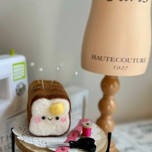 May include: A felt pincushion shaped like a slice of bread with a smiling face and a yellow yolk. The pincushion is on a wooden table with a pink spool of thread, a pair of scissors, and a measuring tape.
