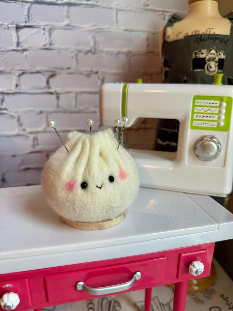 K&ouml;nnte beinhalten: Ein handgefertigtes, cremefarbenes Filz-Kn&ouml;del-Nadelkissen mit einem l&auml;chelnden Gesicht und rosa Wangen. Perlenkopf-Nadeln sind in die Oberseite gesteckt. Das Nadelkissen steht auf einem kleinen Holzsockel neben einer wei&szlig;en N&auml;hmaschine.