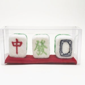 May include: Three white felt mahjong tiles with green and red embroidered characters. The tiles are displayed in a clear plastic box on a red felt base.