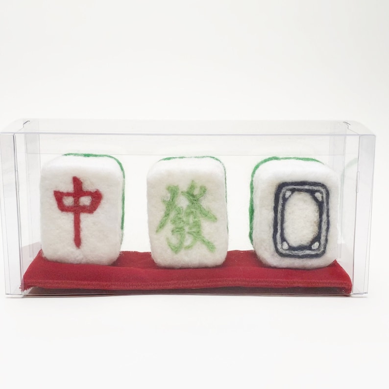 May include: Three white felt mahjong tiles with green and red embroidered characters. The tiles are displayed in a clear plastic box on a red felt base.