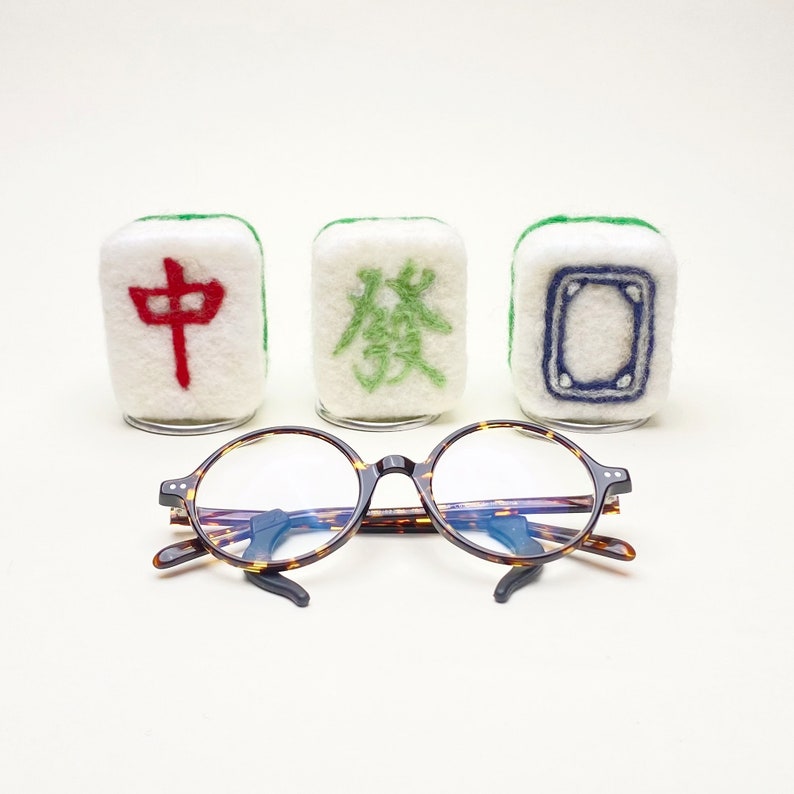 May include: Three white felt mahjong tiles with green and red Chinese characters. The tiles are arranged in a row. A pair of tortoiseshell round glasses are in front of the tiles.
