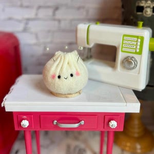 K&ouml;nnte beinhalten: Ein Filzkn&ouml;del mit einem l&auml;chelnden Gesicht und rosa Wangen sitzt auf einem kleinen Holzsockel. Im Hintergrund befindet sich eine wei&szlig;e Spielzeugn&auml;hmaschine mit gr&uuml;nen Akzenten. Der Kn&ouml;del steht auf einem rosa-wei&szlig;en Tisch.