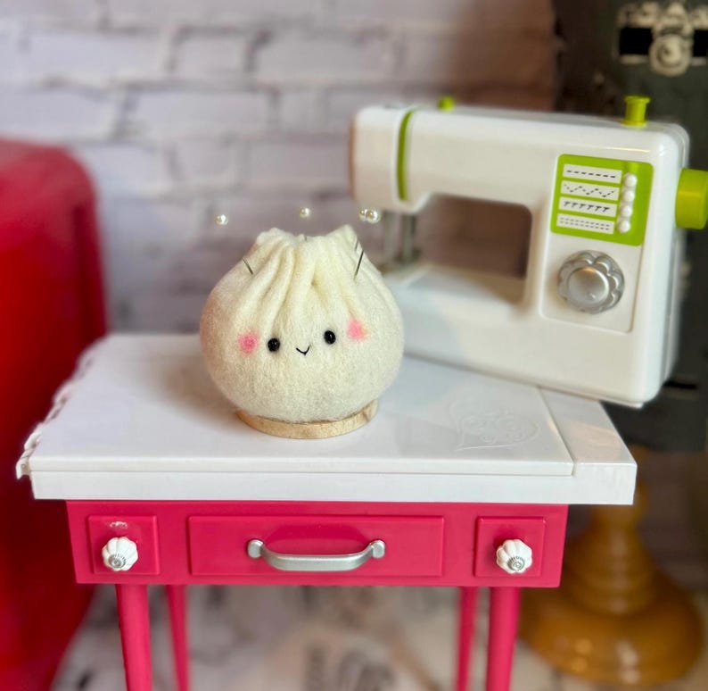 K&ouml;nnte beinhalten: Ein Filzkn&ouml;del mit einem l&auml;chelnden Gesicht und rosa Wangen sitzt auf einem kleinen Holzsockel. Im Hintergrund befindet sich eine wei&szlig;e Spielzeugn&auml;hmaschine mit gr&uuml;nen Akzenten. Der Kn&ouml;del steht auf einem rosa-wei&szlig;en Tisch.