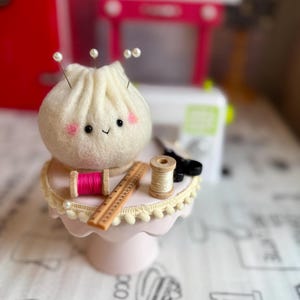 May include: A whimsical felt dumpling character with pink cheeks and pearl pins, atop a pink cake stand. The stand is adorned with sewing notions: pink thread, a ruler, and scissors. A miniature sewing machine and a red refrigerator are in the background.