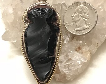 Puede incluir: Un colgante de obsidiana negra con un marco de metal plateado y una moneda de veinticinco centavos para comparar el tamaño. El colgante tiene forma de lágrima y una superficie texturizada. El marco tiene un borde con cuentas.