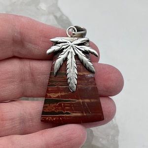 Puede incluir: Un colgante de plata con un diseño de hoja de marihuana estilizado, colocado sobre una piedra rayada roja y marrón.