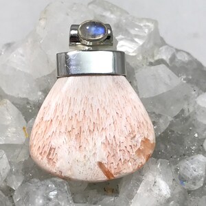 Op de afbeelding: Een zilveren hanger met een witte en roze edelsteen en een maansteen accent. De hanger ligt op een bed van witte kristallen.