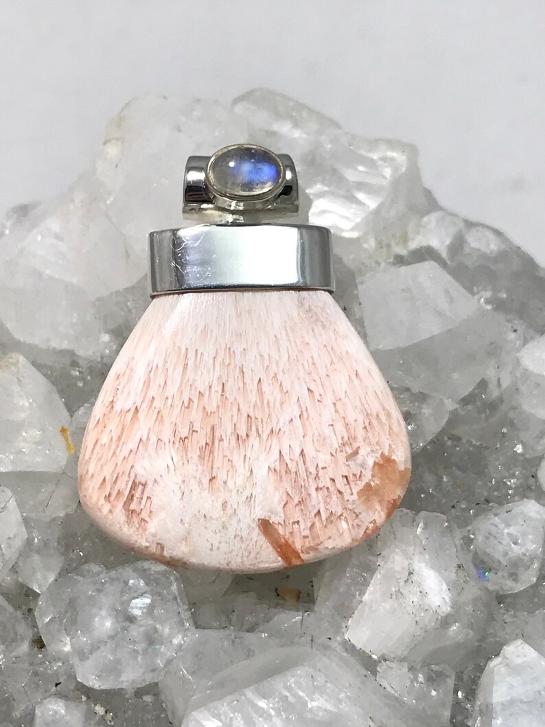 Op de afbeelding: Een zilveren hanger met een witte en roze edelsteen en een maansteen accent. De hanger ligt op een bed van witte kristallen.