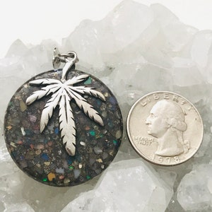 May include: A silver marijuana leaf pendant with a dark gray, round, resin base filled with colorful glitter. The pendant is next to a US quarter for size comparison.