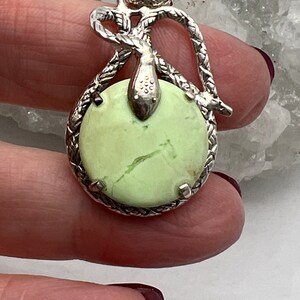 May include: A silver pendant with a green serpentine stone set in a rope-like design. The pendant features a snake design with a knotted tail.