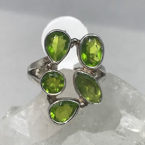 Può includere: Un anello d'argento con cinque pietre di peridoto di varie forme. Le pietre di peridoto sono di un verde brillante, incastonate in una fascia d'argento. Il design dell'anello è un gruppo di gemme, che crea un gioiello unico.