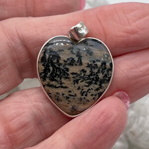 May include: Heart-shaped pendant with a silver-colored bezel. The stone has a natural pattern of black and tan, resembling a landscape. The pendant is held in a hand, showcasing the size and detail of the jewelry.
