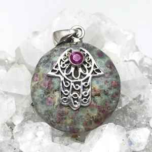 May include: A silver hamsa hand charm pendant with a ruby gemstone set in the centre. The pendant is made of a green and pink stone with a speckled pattern.