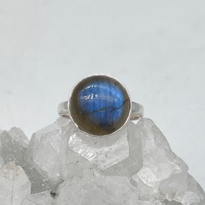May include: A silver ring featuring a large, round labradorite stone. The stone has a blue and gray iridescent sheen. The ring is set on a white, crystalline surface.