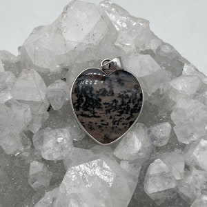 May include: Heart-shaped pendant with a silver-colored border. The pendant features a natural stone with a pattern of black and brown hues. The pendant is displayed on a bed of clear, crystalline formations.