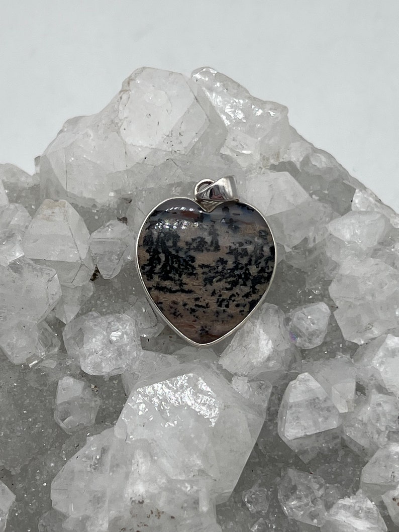 May include: Heart-shaped pendant with a silver-colored border. The pendant features a natural stone with a pattern of black and brown hues. The pendant is displayed on a bed of clear, crystalline formations.