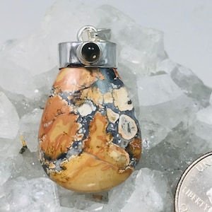 May include: A teardrop-shaped pendant with a silver bail and a small, dark stone at the top. The pendant features a mix of tan, brown, gray, and white colors. The stone has a unique, natural pattern. The pendant is displayed on a white, crystalline surface.