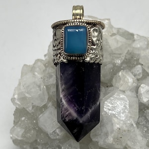 May include: A silver pendant featuring a rectangular, light blue stone in an ornate setting. The pendant's base is a dark purple crystal with white veining. The pendant is displayed on a white, crystalline surface.