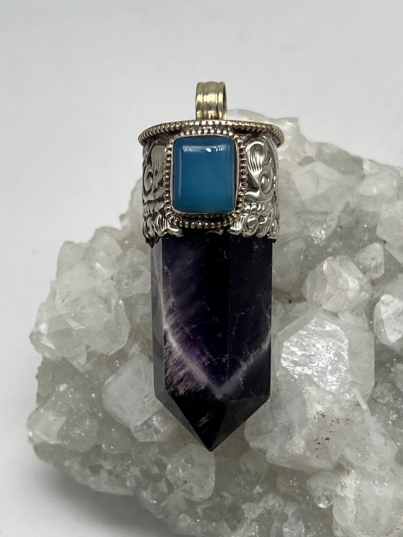 May include: A silver pendant featuring a rectangular, light blue stone in an ornate setting. The pendant's base is a dark purple crystal with white veining. The pendant is displayed on a white, crystalline surface.