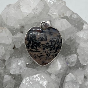 May include: Heart-shaped pendant with a silver-colored bail. The pendant features a natural stone with a pattern of black and brown inclusions. The stone is set in a silver-colored bezel.