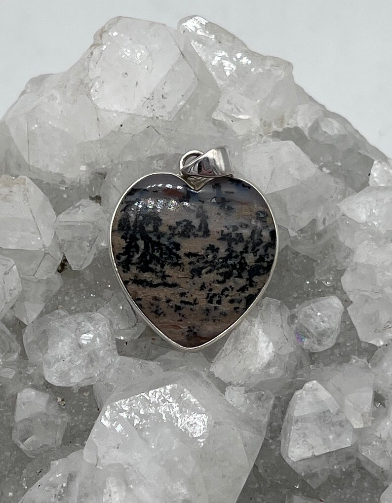 May include: Heart-shaped pendant with a silver-colored bail. The pendant features a natural stone with a pattern of black and brown inclusions. The stone is set in a silver-colored bezel.