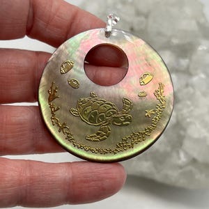 Peut inclure: Pendentif rond avec une finition en nacre, représentant une tortue de mer, des poissons et des coraux dorés. Le pendentif est doté d'une bélière argentée pour l'accrocher. La surface irisée reflète des teintes roses, vertes et dorées.