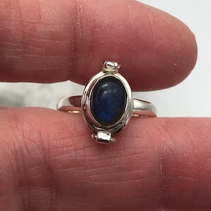 Può includere: Un anello d'argento con una pietra preziosa ovale blu scuro incastonata in una lunetta decorativa. L'anello ha una parte superiore incernierata e un piccolo elemento decorativo nella parte superiore della lunetta. L'anello è tenuto tra le dita.