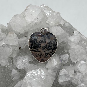 May include: Heart-shaped pendant with a silver-colored border. The pendant features a unique pattern of black and brown inclusions within the stone. The pendant is displayed against a backdrop of clear, crystalline formations.
