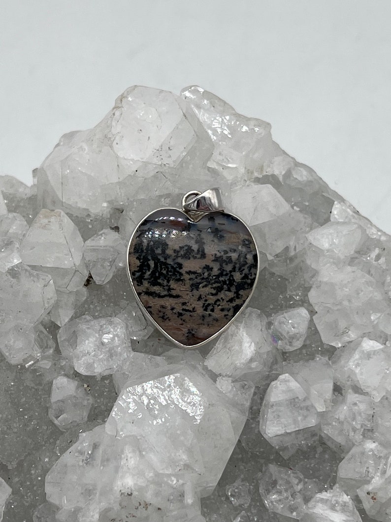 May include: Heart-shaped pendant with a silver-colored border. The pendant features a unique pattern of black and brown inclusions within the stone. The pendant is displayed against a backdrop of clear, crystalline formations.