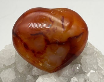 Puede incluir: Una piedra de cornalina pulida en forma de corazón. La piedra es de un color naranja rojizo cálido con vetas y marcas más oscuras. Descansa sobre un lecho de cuarzo cristalino blanco.