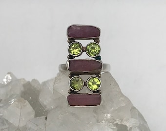 May include: A silver ring featuring three rectangular pink stones and two green stones. The ring is displayed on a bed of white crystals.